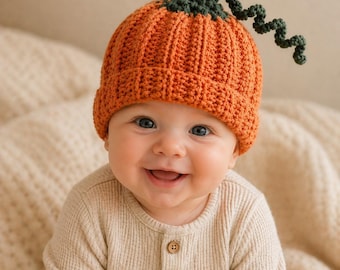 May include: A smiling baby wearing an orange crochet pumpkin hat with a green stem and curly vine. The baby is dressed in a beige waffle-knit onesie with wooden buttons. The background is a soft, neutral color.