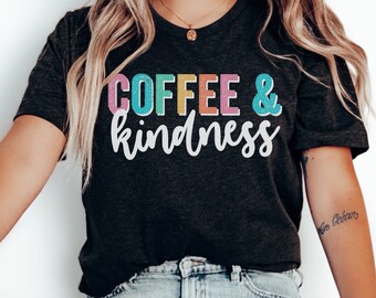 May include: Dark gray t-shirt with the words "COFFEE & kindness" in colorful, playful lettering. The shirt is paired with light wash jeans and white slip-on shoes. The person is sitting on a wooden stool.