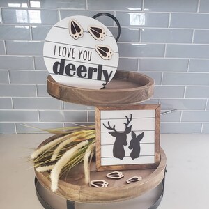 Puede incluir: Una bandeja de madera de dos niveles que presenta un letrero redondo con el texto I LOVE YOU deerly y tres pequeñas huellas de pezuñas, un letrero cuadrado con la silueta negra de dos ciervos y un manojo de tallos de hierba seca con más huellas.
