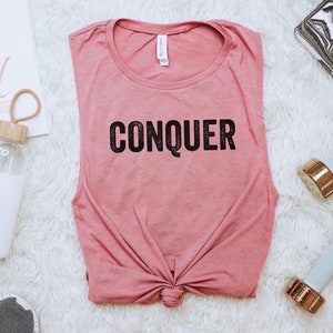 May include: A dusty rose tank top with the word "CONQUER" printed in black. The image includes a water bottle, dumbbells, and a motivational print that says "You Go Girl". The items are arranged on a white, fluffy surface.