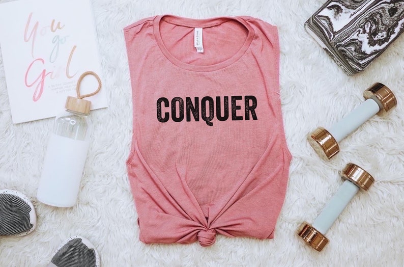 May include: A dusty rose tank top with the word "CONQUER" printed in black. The image includes a water bottle, dumbbells, and a motivational print that says "You Go Girl". The items are arranged on a white, fluffy surface.