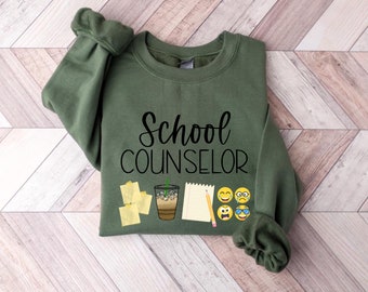 May include: A khaki green sweatshirt featuring the text "School Counselor" in black. The design includes a coffee, sticky notes, a pencil, a notepad, and emoji faces.