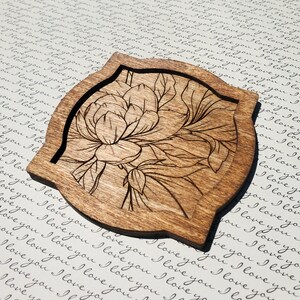 Puede incluir: Un posavasos de madera con un diseño de flor grabado con láser. El posavasos es marrón y tiene una forma redondeada con un borde biselado.