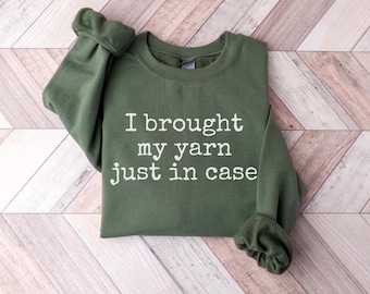 May include: An olive green crewneck sweatshirt featuring the white text "I brought my yarn just in case" printed across the chest. The garment has long sleeves with ribbed cuffs and a ribbed neckline, showcasing a simple and functional design.