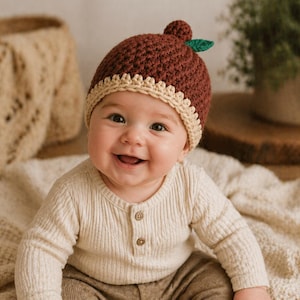 Könnte beinhalten: Ein lächelndes Baby trägt einen handgefertigten braunen Eichelhut mit beigem Rand und einem kleinen grünen Blatt. Das Baby ist in einem cremefarbenen Langarmshirt und einer braunen Hose gekleidet und sitzt auf einer cremefarbenen Decke.