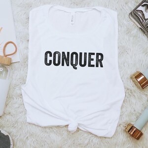 May include: White sleeveless workout top with the word "CONQUER" in black. The image includes a water bottle, two dumbbells with gold-colored ends, and a notebook with the text "You Go Girl".