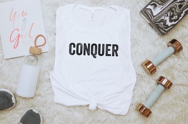 May include: White sleeveless workout top with the word "CONQUER" in black. The image includes a water bottle, two dumbbells with gold-colored ends, and a notebook with the text "You Go Girl".