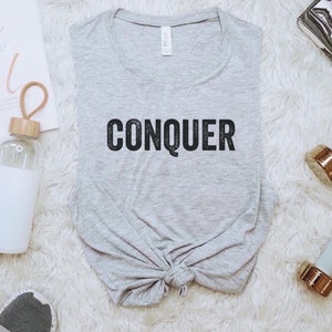 May include: A light gray t-shirt with the word "CONQUER" in black block letters. The shirt is knotted at the bottom. The image also includes a water bottle, dumbbells, and a motivational print that says "You go Girl".