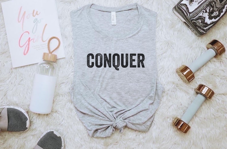 May include: A light gray t-shirt with the word "CONQUER" in black block letters. The shirt is knotted at the bottom. The image also includes a water bottle, dumbbells, and a motivational print that says "You go Girl".