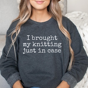 May include: A dark charcoal gray crewneck sweatshirt featuring the white text "I brought my knitting just in case" printed across the chest. The garment has long sleeves with ribbed cuffs and a ribbed neckline, providing a standard fit.