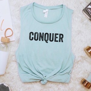May include: A light blue tank top with the word "CONQUER" in black lettering, tied at the waist, is surrounded by fitness accessories. A water bottle, dumbbells, and a marble-patterned block are also present. A motivational print reads "You Go Girl."