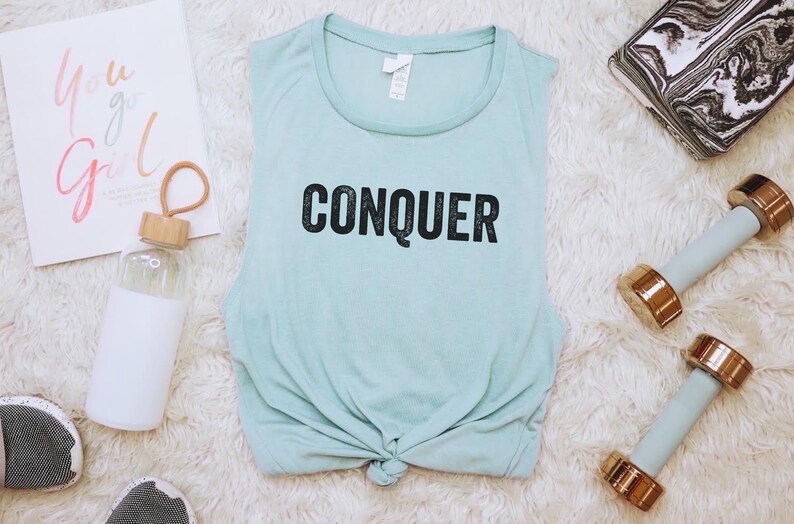 May include: A light blue tank top with the word "CONQUER" in black lettering, tied at the waist, is surrounded by fitness accessories. A water bottle, dumbbells, and a marble-patterned block are also present. A motivational print reads "You Go Girl."