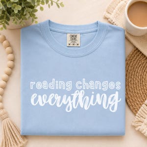 May include: A light blue crew neck t-shirt with the white text reading changes everything printed on the front. The shirt features a Comfort Colors brand label at the inner neckline, indicating a size M, and has a standard short-sleeve construction.