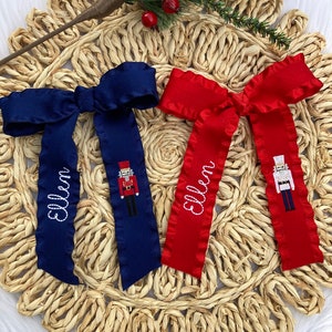 May include: Two personalized Christmas bows with a nutcracker design. One bow is red and the other is blue. The bows are embroidered with the names "Ellen" and "Elim".