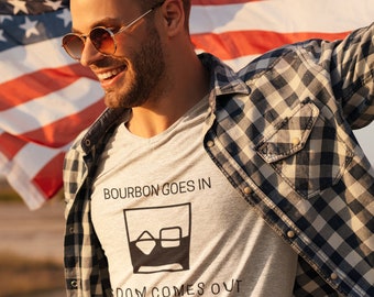 May include: A man wearing a grey t-shirt with the text "Bourbon Goes In Freedom Comes Out" and a glass of bourbon with ice cubes graphic. He is holding an American flag in his right hand and smiling.