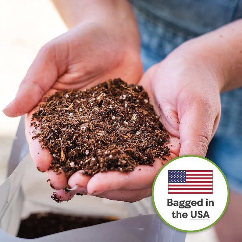 May include: Close-up of a person's hands holding a handful of dark brown potting soil. The soil is a mix of dark brown compost and white perlite. A white bag with the text "Bagged in the USA" is visible in the background.