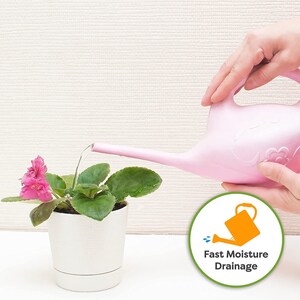 May include: A pink watering can with a long spout is pouring water onto a potted plant with pink flowers. The plant is in a white pot with a drainage hole. The watering can has a handle and a decorative design. A green circle with the text "Fast Moisture Drainage" and a small orange watering can icon is in the lower right corner of the image.