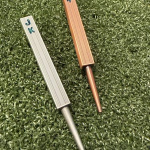May include: Two metal golf divot repair tools with a rectangular handle featuring a textured grid pattern and a tapered spike. One tool is silver-toned and the other is copper-toned. Both handles are engraved with the letters JK in blue.