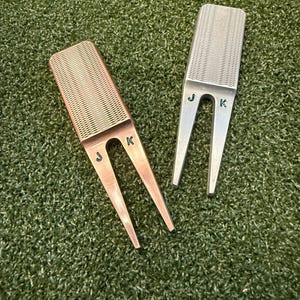 May include: Two golf divot repair tools. One is bronze-colored, the other is silver. Both have a rectangular top with a grid pattern and two prongs. The bronze tool has the letter "J" and "K" on it. The silver tool has the letter "J" and "K" on it.
