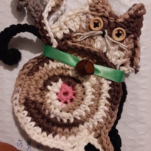 Può includere: Applique a forma di gatto all'uncinetto fatto a mano nei toni del marrone, crema e nero, con occhi a bottone e un collare di nastro verde. Il gatto ha un dettaglio floreale rosa e una piccola campana. Un oggetto artigianale stravagante.