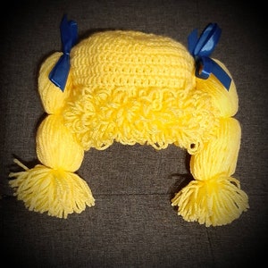 May include: A yellow crocheted wig with blue ribbon bows. The wig has long, curly strands of yarn.