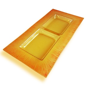 Puede incluir: Un plato de servir rectangular de vidrio con dos compartimentos cuadrados. El plato tiene una base amarilla con un patrón de sol rojo y naranja alrededor de los bordes.