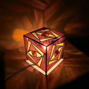 以下が含まれることがあります： 3つの黄色い目を特徴とする幾何学模様のステンドグラス製の立方体ランプ。ランプは点灯しており、温かい光を放っています。