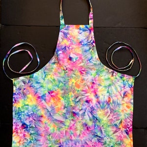 May include: A colorful tie-dye apron with a vibrant mix of pink, blue, yellow, and green hues. The apron features a neck loop and waist ties, perfect for cooking or crafting. The fabric has a crinkled texture.