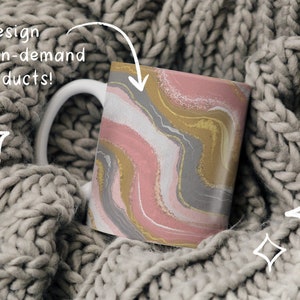 May include: A white ceramic mug with a pink, gold, and gray abstract design. The mug is sitting on a gray knitted blanket.