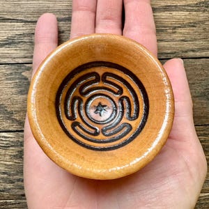 May include: A small, round, ceramic bowl with a brown glaze. The bowl has a black and white design of a labyrinth with a star in the center.