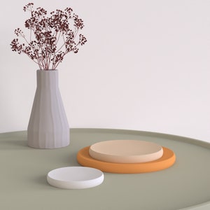Könnte beinhalten: Ein rundes Tablett in Hellgrün mit zwei runden Plattformen. Die obere Plattform ist hellbraun und die untere Plattform ist orange. Eine weiße Vase mit getrockneten Blumen befindet sich auf der linken Seite des Tabletts.