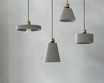 May include: Four gray concrete pendant lights with brass-colored bulb holders hang from black cords. The shades have different shapes: a flat disc, a cone, a truncated cone, and a cylinder. The background is a light gray wall.
