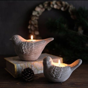 Puede incluir: Dos candelabros de pájaro de hormigón gris con velas de té encendidas en su interior. Los candelabros están sobre un libro con una piña delante.