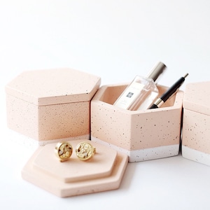 May include: Three hexagonal pink and white speckled concrete boxes with a white stripe around the top. The middle box has a clear glass bottle and a black pen inside. The box on the left has two gold earrings with a speckled design on top.