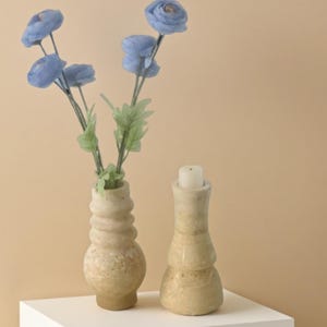May include: A still life featuring a light-coloured, textured vase holding blue artificial flowers. Beside it, a matching candle holder with a white candle sits on a white surface against a neutral background.