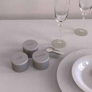 May include: Three grey ceramic containers labelled "salt", "pepper", and "sugar" on a white table setting with two champagne glasses and a plate.