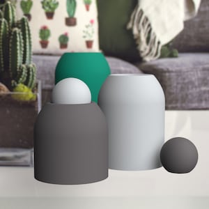 Op de afbeelding: Drie decoratieve containers, een grijs, een wit en een groen, met een witte bol in de grijze container en een grijze bol op de tafel.