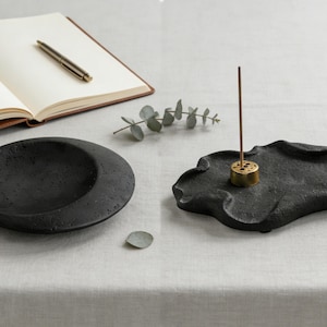 Puede incluir: Un plato redondo negro texturizado y un porta incienso negro de forma orgánica con un porta incienso dorado. También hay un cuaderno con un bolígrafo y una ramita de vegetación.