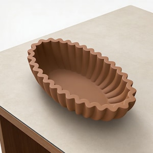 Può includere: Una ciotola decorativa marrone, di forma ovale, con bordo scanalato. La ciotola è realizzata in un materiale solido e poggia su una superficie di colore chiaro. Il design è semplice ed elegante, adatto a vari stili di arredamento.
