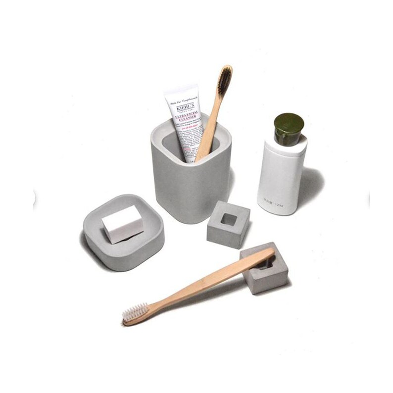 以下が含まれることがあります： グレーのコンクリート製のバスルームアクセサリー4点セット。セットには、歯ブラシホルダー、石鹸置き、タンブラー、ディスペンサーが含まれています。ディスペンサーは白で、緑色の蓋が付いています。歯ブラシホルダーには、歯ブラシを入れるための小さな穴があります。石鹸置きには、石鹸を置くための小さな窪みがあります。タンブラーは背が高く、円筒形です。ディスペンサーは短く、円筒形です。