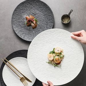 Könnte beinhalten: Drei strukturierte Keramikteller, einer schwarz und zwei weiß, mit einer strukturierten Oberfläche. Die Teller sind auf einer grauen Oberfläche angeordnet. Ein Teller hat eine kleine Portion Tintenfisch und eine Birnenspalte darauf. Ein weiterer Teller hat eine Portion Jakobsmuscheln und Grün darauf.