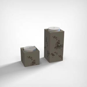 Op de afbeelding: Twee grijze betonnen kandelaars van verschillende hoogtes. Elk heeft een ronde witte kaars erin. Het beton heeft een verweerde, getextureerde uitstraling met donkere vlekken, wat het een rustieke uitstraling geeft. De kandelaars zijn vierkant en modern.
