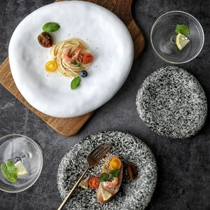 May include: A selection of contemporary tableware. A large white organic-shaped plate holds pasta and shrimp. A grey speckled plate with bread, and small glass bowls with lemon and mint are also shown.