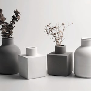 Op de afbeelding: Vier minimalistische vazen in grijstinten en wit. Twee vazen bevatten gedroogde planten. De vazen hebben verschillende vormen, waaronder een afgeronde fles, een vierkant blok en een cilindrisch ontwerp. Voor interieurdecoratie.