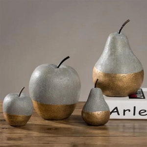 May include: Four decorative fruit sculptures: two pears and two apples. The top halves are gray, and the bottom halves are gold. Each fruit has a black stem. The sculptures are arranged on a wooden surface.