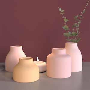 Può includere: Quattro vasi in ceramica di diverse dimensioni e colori. Due sono rosa chiaro, uno è giallo pallido e uno è rosa scuro. I vasi sono disposti su un tavolo con una piccola candela bianca davanti a loro.