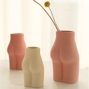 May include: Three ceramic vases shaped like a torso. One is beige, and two are pink. One pink vase holds a dried flower. The vases have a matte finish and are arranged on a white surface.