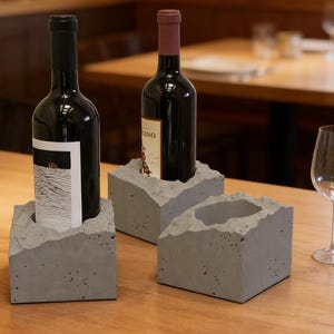 Puede incluir: Tres soportes de hormigón gris para botellas de vino con un acabado rugoso y texturizado. Dos soportes contienen botellas de vino oscuras y un tercero está vacío. Una copa de vino está sobre la mesa de madera. Los soportes son angulares.