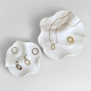 Pode incluir: Duas bandejas brancas para joias com formato orgânico com várias joias douradas e prateadas. As bandejas contêm um colar de ouro com um pingente quadrado, um anel de ouro e outros anéis e brincos.