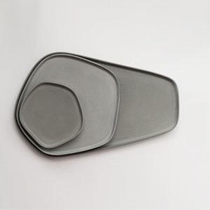 Puede incluir: Tres bandejas de servir de cerámica gris de diferentes tamaños, anidadas entre sí. Las bandejas tienen una forma orgánica y libre.